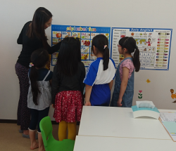 壁にかけたアルファベットとフォニックスの表を見る講師と子供達